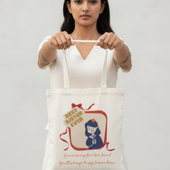 A white canvas tote bag hanging from a black door handle on a white paneled door. The bag features a graphic illustration of a girl in a blue uniform with a red ribbon, next to a sign that reads 'BEST SISTER EVER', and cursive text below that says 'You were my first friend You'll always be my forever home'.