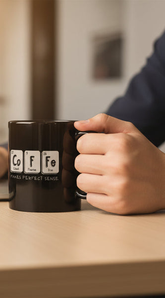 Two black mugs with periodic table elements spelling 'Coffee' on a beige background with colorful candies.